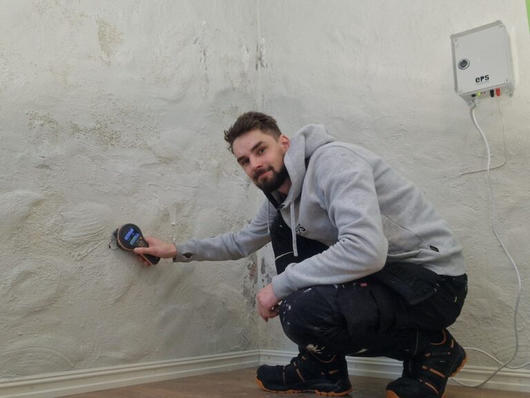 En hyggelig medarbeider viser hvordan han måler fukt på veggen i et kjellerrom. Han smiler imøtekommende til kamera.